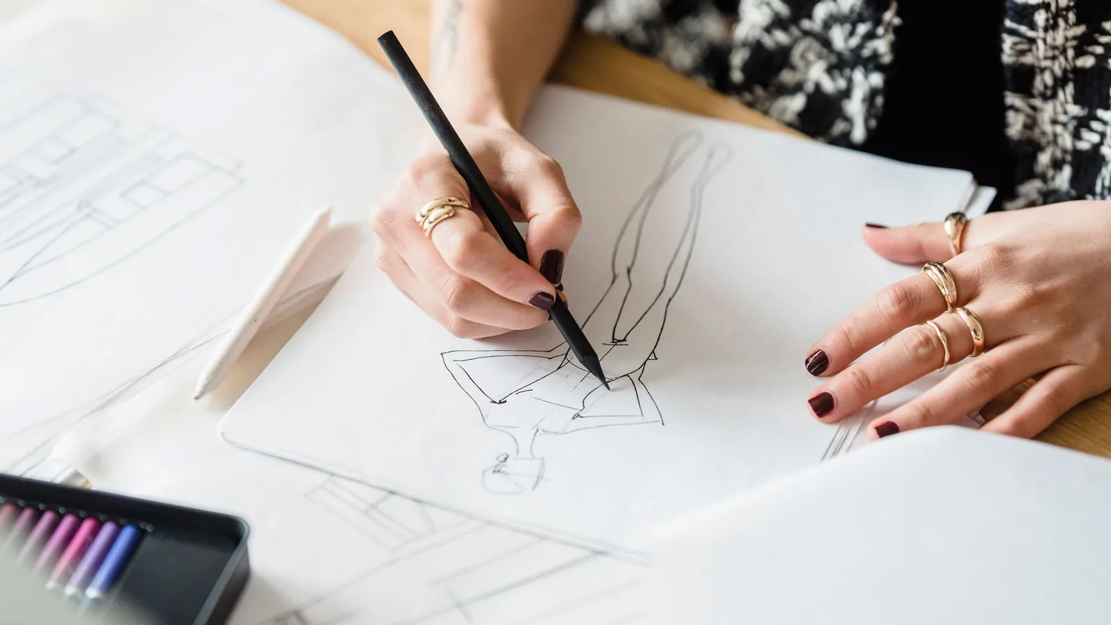 Mão desenhando o rascunho das telas no papel, ao lado das canetas